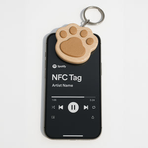 木製貓掌造型鑰匙圈放置於手機上方,呈現 NFC 感應使用情境。鑰匙圈以淺色木材製作,中央嵌有較深色木片,搭配金屬鑰匙圈。手機螢幕顯示音樂播放介面,可見 Spotify 標誌、「NFC Tag」、「Artist Name」以及播放控制按鈕,突顯商品結合 NFC 功能的特色。