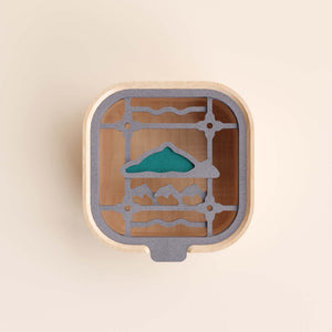 宜蘭窗花物語收納盒系列單品展示圖，盒身以淺色木材製作，搭配灰色內層盒蓋。盒蓋圖樣以窗花框架為基礎，中央結合綠色山形、下方魚群與波浪元素，呈現宜蘭山海景致的在地意象。畫面採正上方視角，清楚展現盒蓋的色彩、圖樣與層次細節。